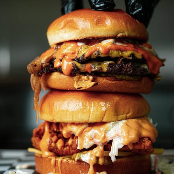 A towering Slaw Burger with two beef patties, fried chicken, cheese, and sauces dripping.