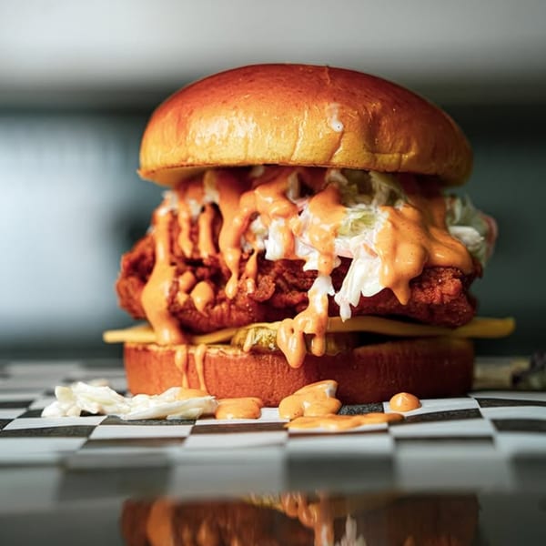 A Slaw Burger with crispy fried chicken, coleslaw, cheese, and orange sauce on a checkered surface.