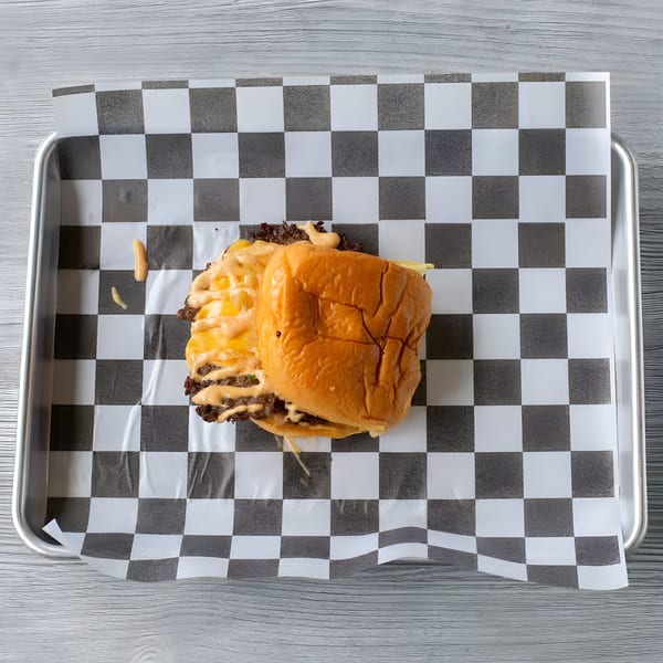 A Slaw Burger with melted cheese and sauce on checkered paper in a metal tray.