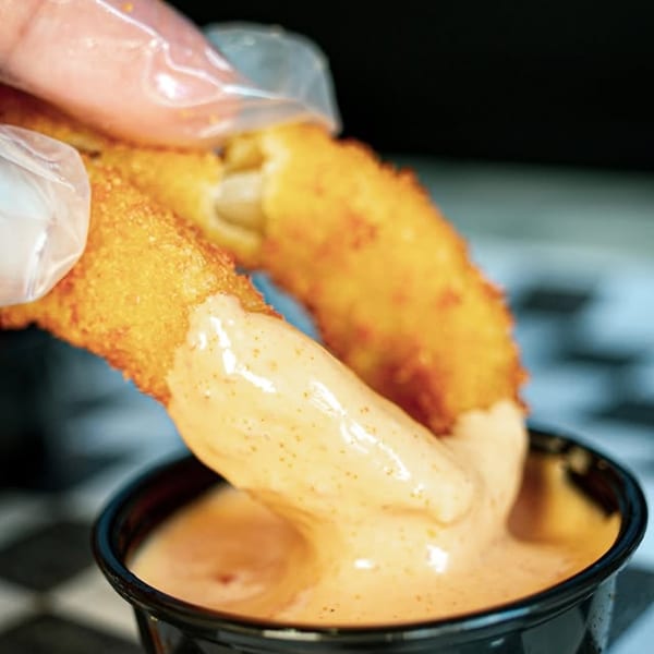A gloved hand dips a golden-brown fried mozzarella stick into a cup of creamy orange sauce.