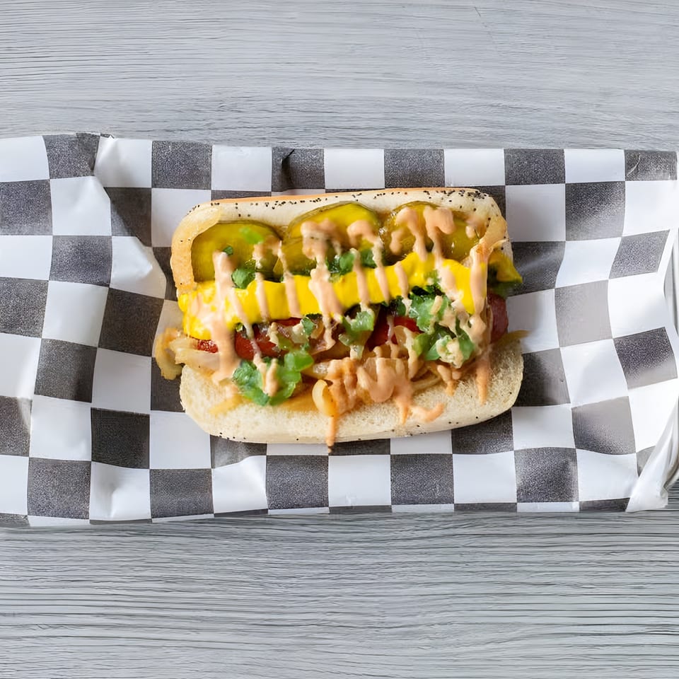 Slaw Burger's hot dog with pickles, mustard, and sauce on checkered paper.