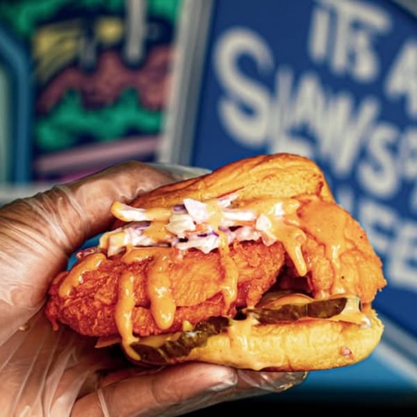 A gloved hand holds a Slaw Burger with fried chicken, slaw, pickles, and sauce on a bun.