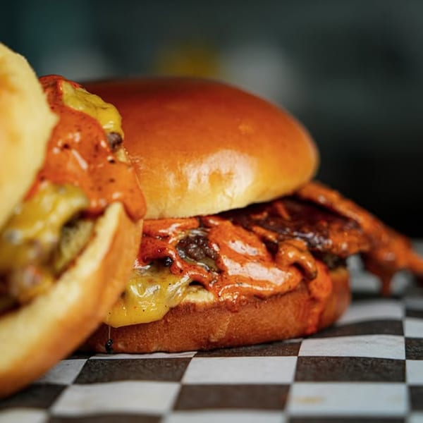 Two Slaw Burger cheeseburgers with orange sauce and melted cheese on a checkered paper.