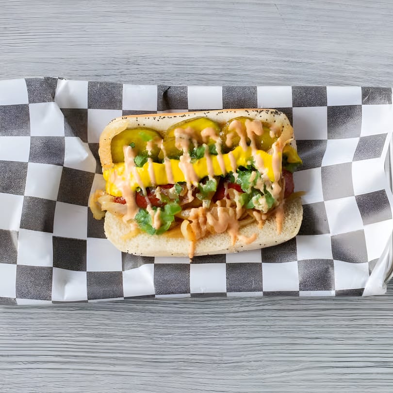 A Slaw Burger hot dog with pickles, mustard, and sauce on checkered paper.