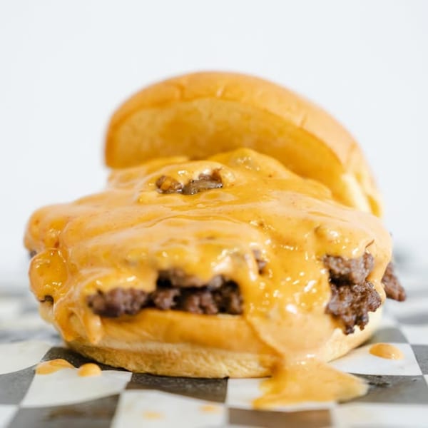 A Slaw Burger with two patties, covered in melted cheese and sauce, on checkered paper.