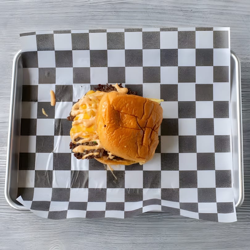 A Slaw Burger with melted cheese and sauce on checkered paper in a metal tray.