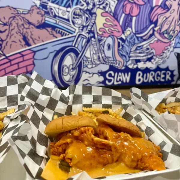 Two Slaw Burger chicken sandwiches with fries in checkered paper, against a colorful mural.