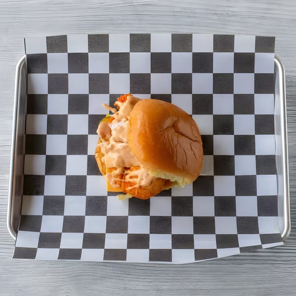 A Slaw Burger with sauce on a checkered paper liner in a metal tray.