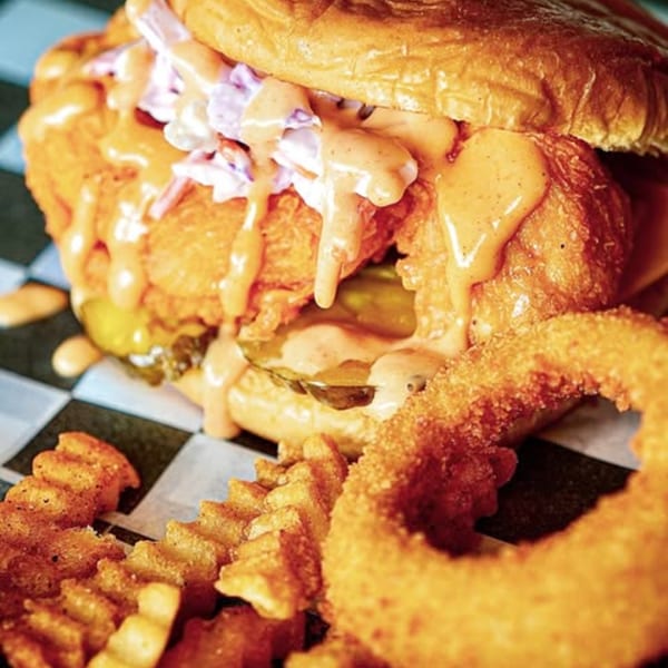 A Slaw Burger with sauce, slaw, pickles, crinkle fries, and an onion ring on checkered paper.